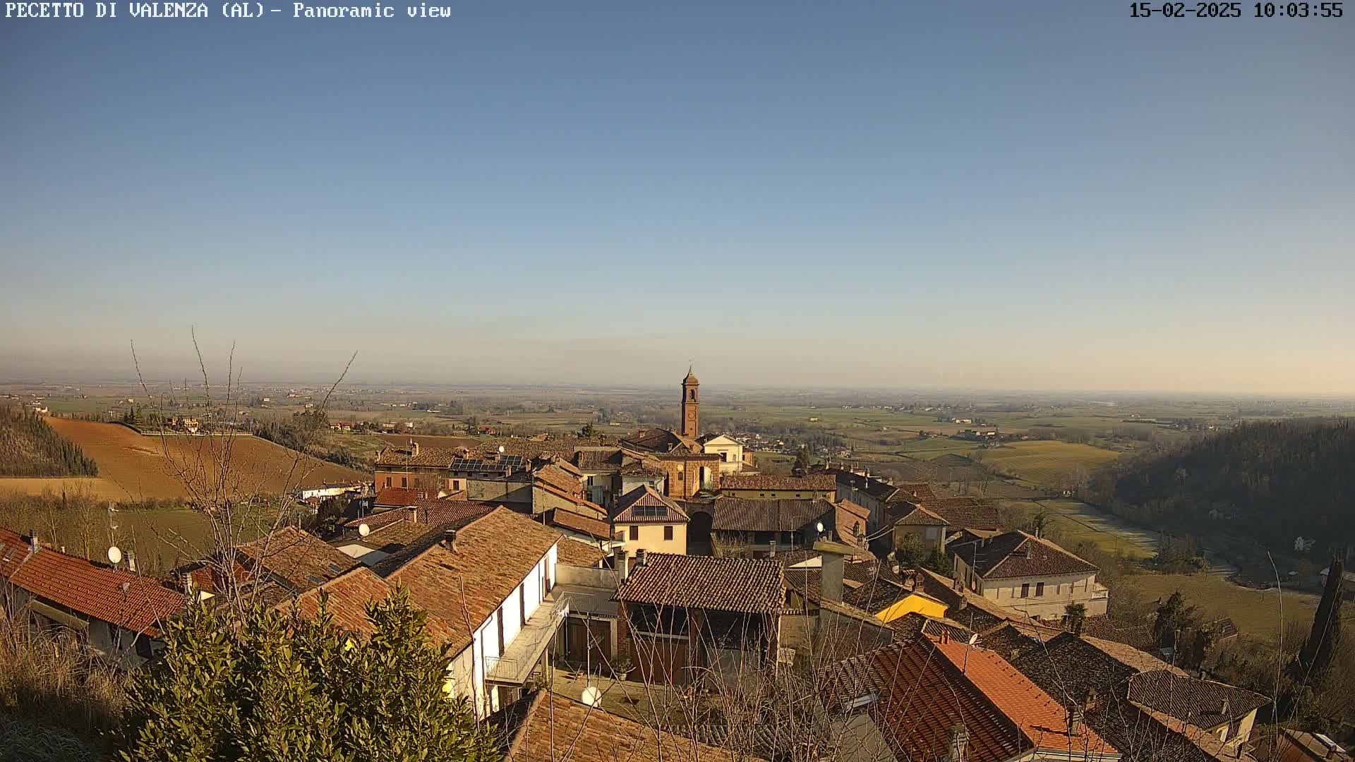 A panoramic view of a village with terracotta-roofed houses, under a clear blue sky, situated on a hillside overlooking a vast, flat agricultural landscape.