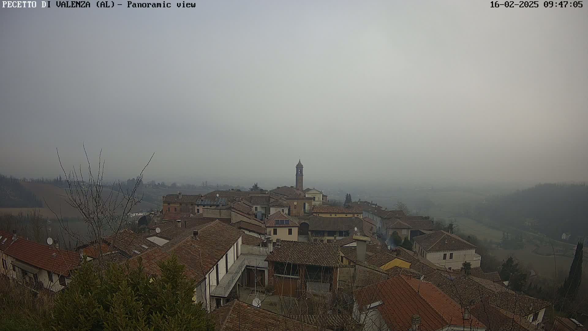 A hazy, overcast view of a village with brown-tiled roofs and a tall bell tower, nestled amongst rolling hills.