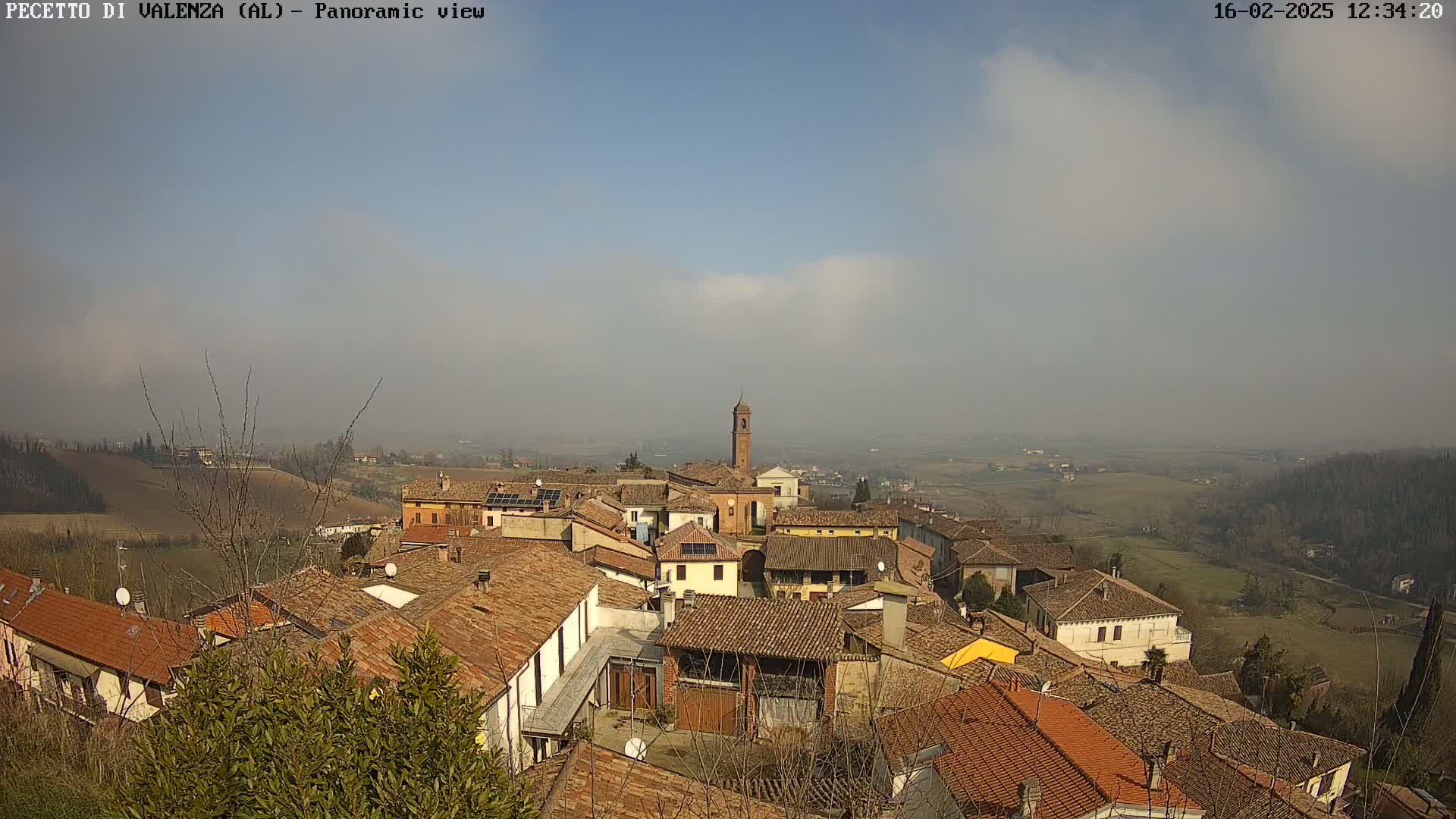 A hazy, sunny day reveals a panoramic view of a small town nestled in rolling hills, with terracotta rooftops dominating the scene and a bell tower rising prominently from the center.