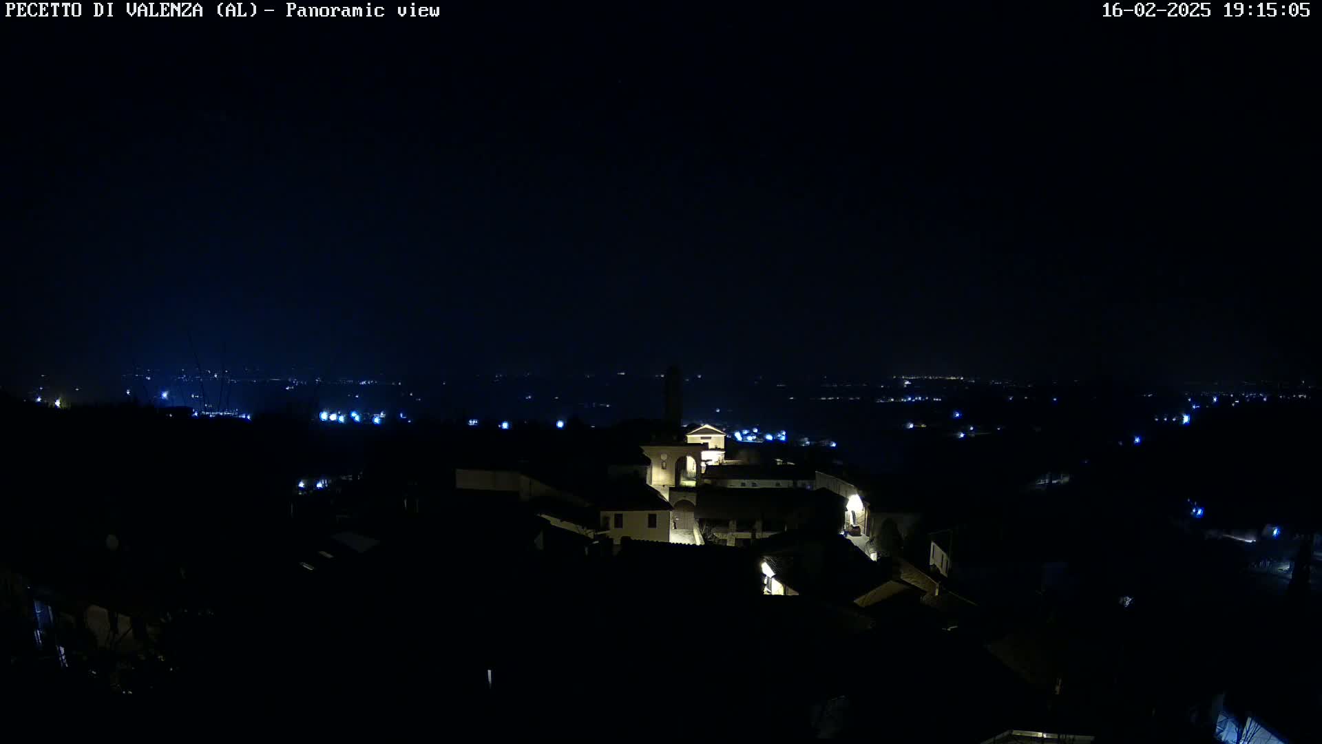 A nighttime panoramic view shows a small town with dimly lit buildings and a few distant lights from other settlements under a dark, clear sky.