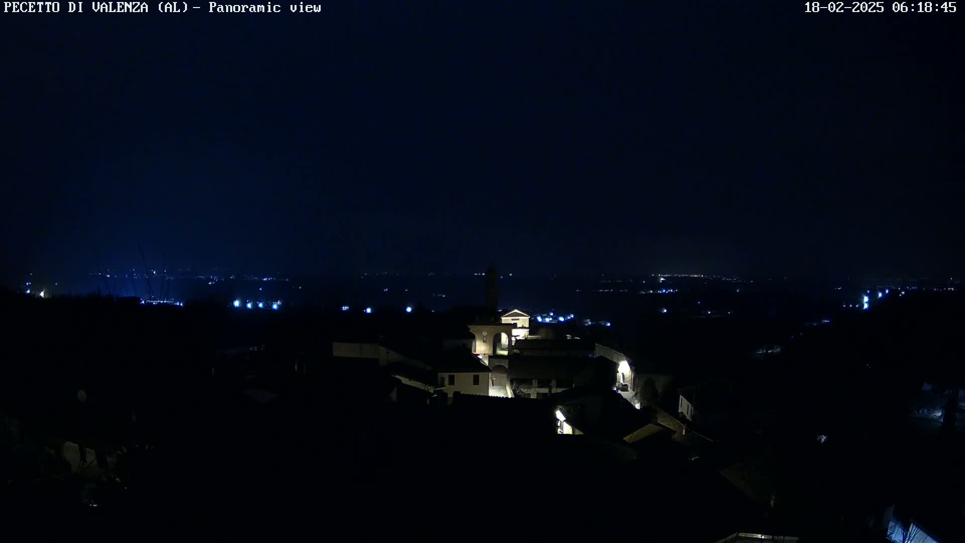 A nighttime panoramic view shows a small village illuminated with scattered lights, overlooking a dark, distant landscape under a clear, dark sky.