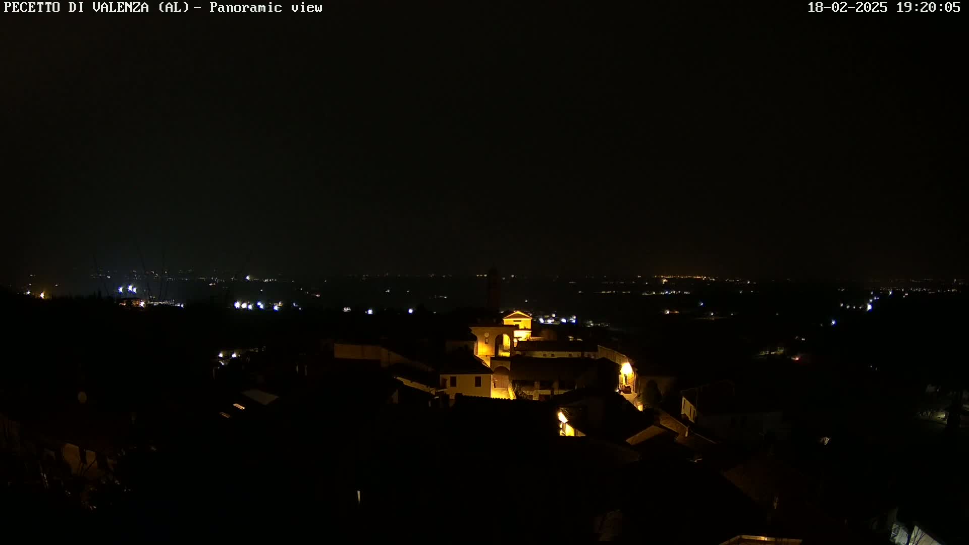 A nighttime panoramic view shows a small, illuminated town nestled in a dark landscape, with more distant lights visible on the horizon.