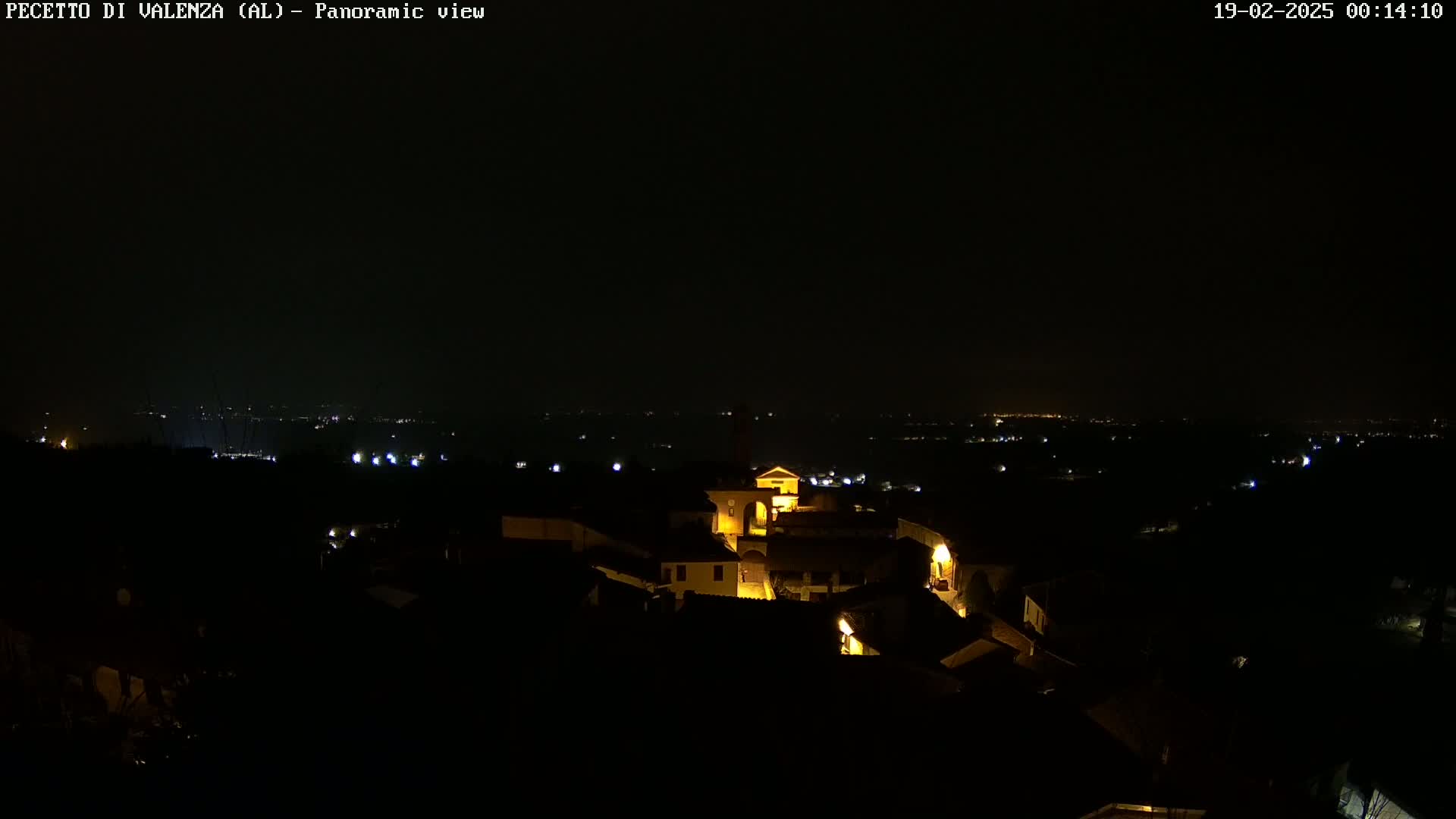 A nighttime panoramic view shows a small, dimly lit town nestled in a dark landscape under a clear, black sky.