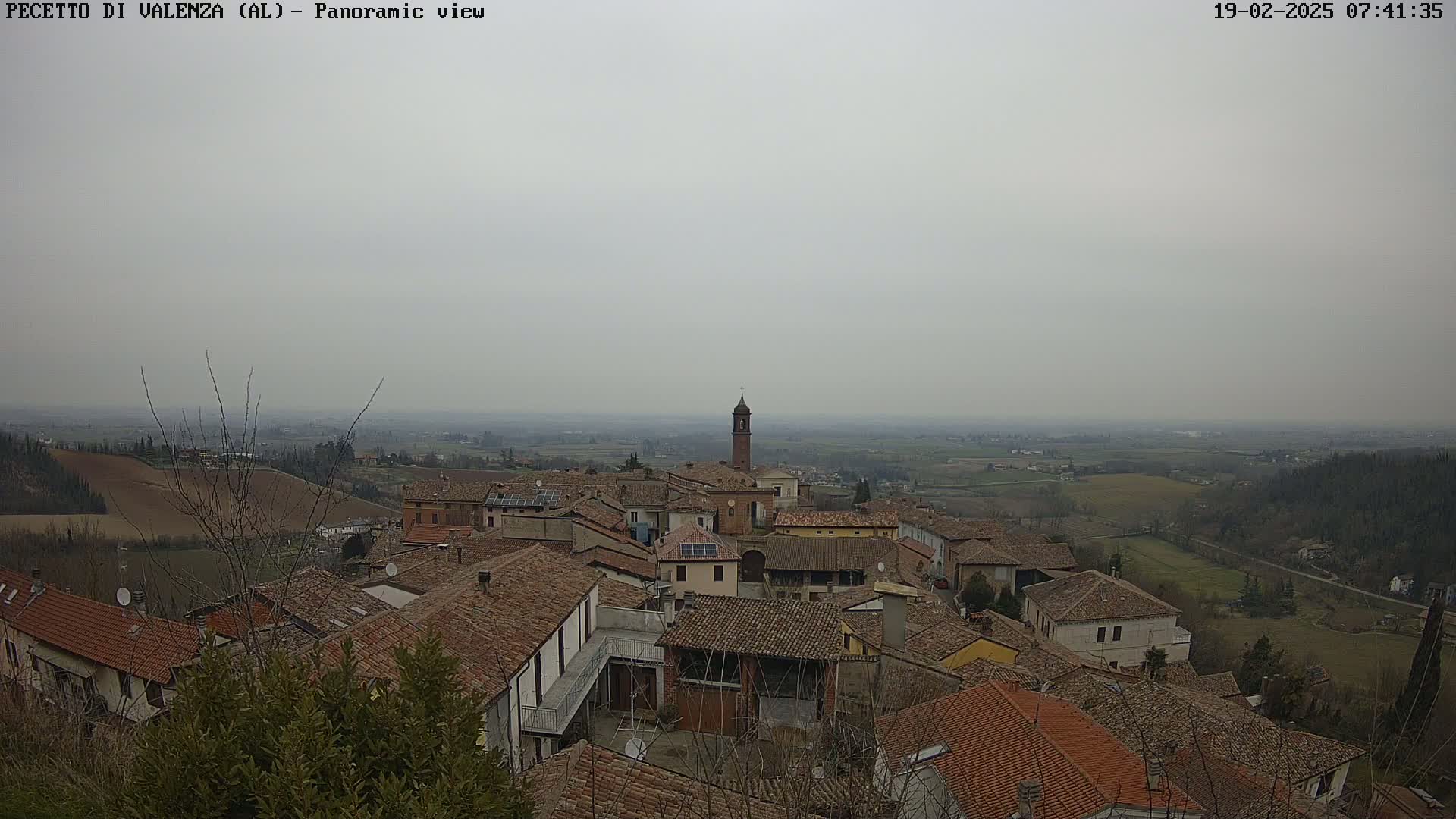 A hazy, overcast view overlooks a small town with terracotta rooftops, a bell tower, and surrounding farmland.
