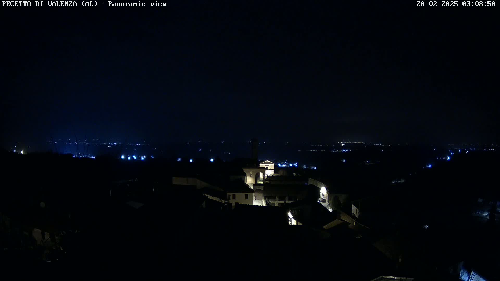 A nighttime panoramic view shows a small, dimly lit town nestled in a dark landscape under a clear, starless sky.