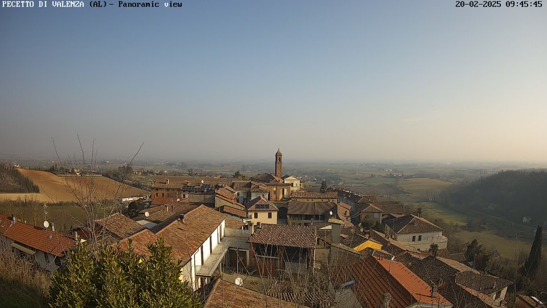 A panoramic view of a small town nestled in a valley on a hazy, sunny day, with terracotta-roofed buildings and a bell tower visible.