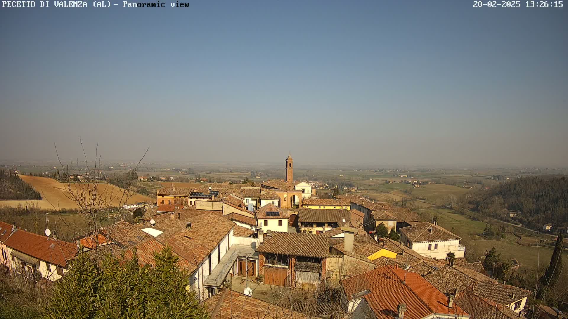 A panoramic view of a small town with terracotta roofs, under a clear, sunny sky, overlooks a flat, agricultural landscape.