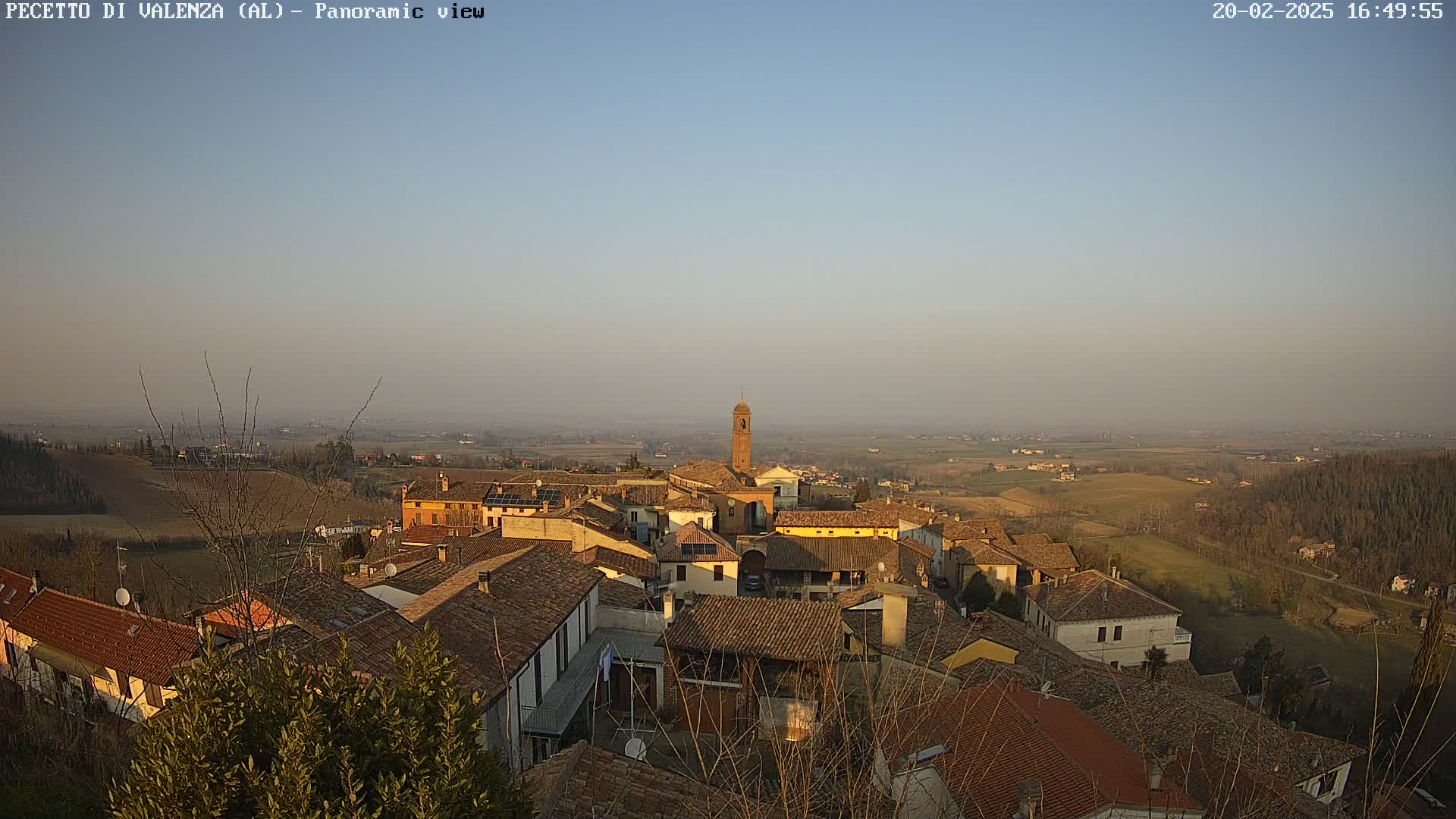 An elevated view of a village with terracotta rooftops, a bell tower, and surrounding farmland under a hazy, clear sky.