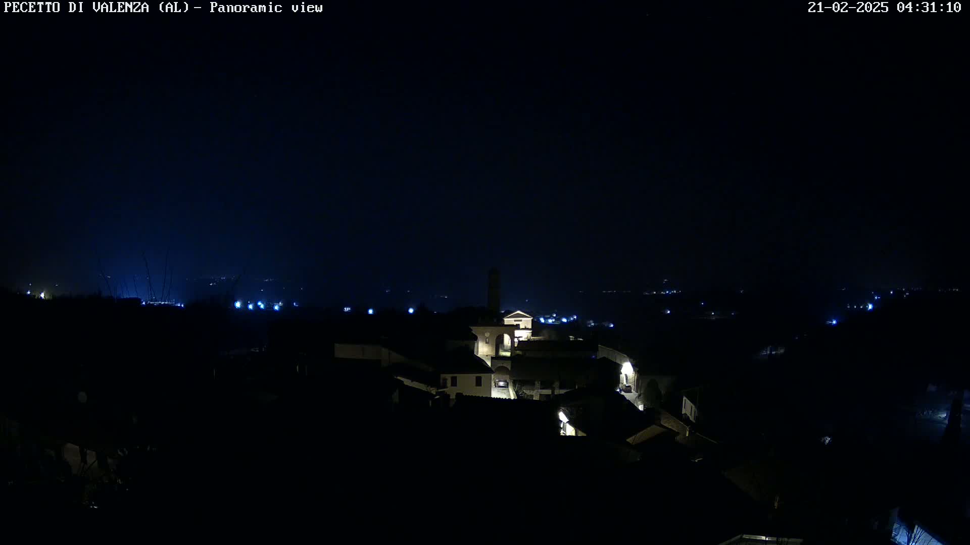 A nighttime panoramic view shows a small, dimly lit town nestled in a dark landscape under a clear, starless sky.