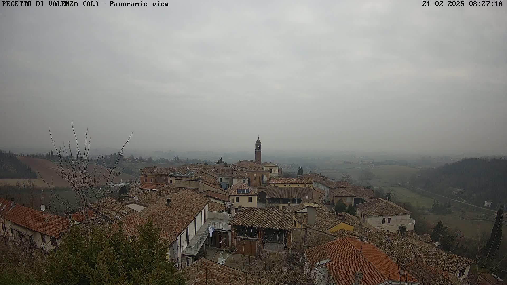 A hazy, overcast day reveals a sprawling village of terracotta-roofed buildings nestled in a gently rolling landscape.