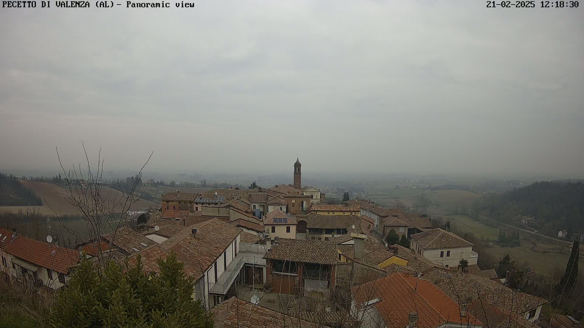 A hazy, overcast day reveals a panoramic view of a small village nestled in a hilly landscape, with numerous terracotta-roofed buildings and a prominent bell tower.