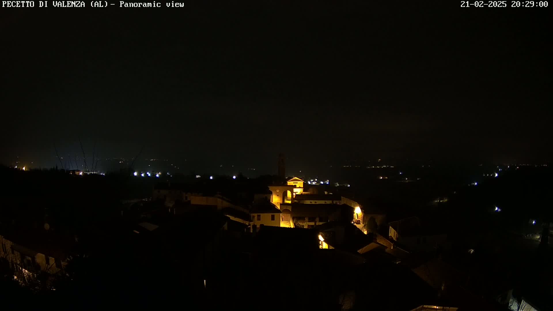 A nighttime panoramic view shows a small, dimly lit town nestled in a dark landscape under a clear, black sky.