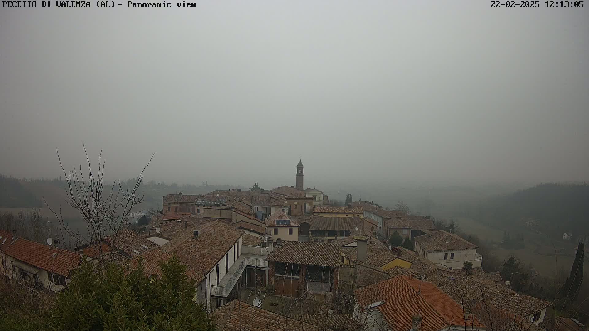 A foggy view of a small town with many brown-roofed buildings, a bell tower, and surrounding hills.