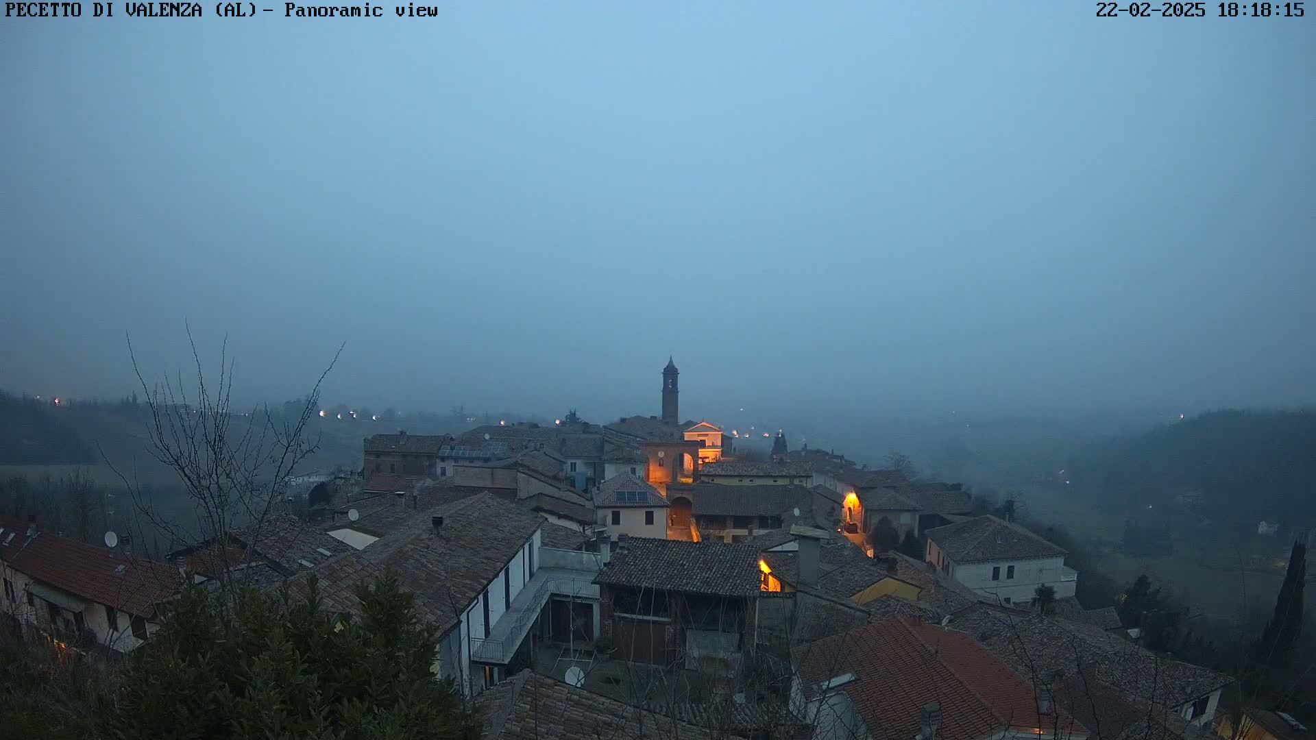 A foggy, twilight view of a small town nestled in a valley, with dimly lit buildings and a church tower visible.