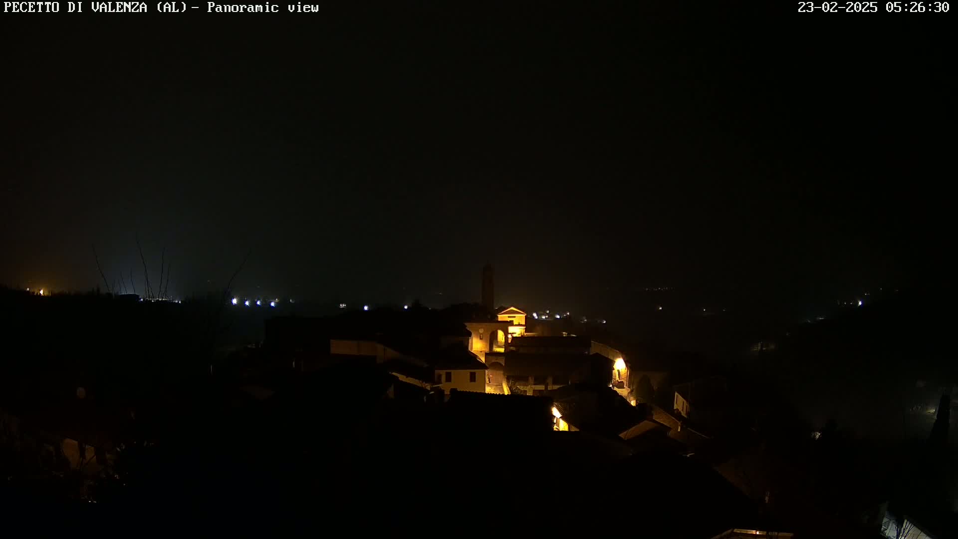 A dimly lit village is visible at night under foggy conditions, with some buildings illuminated.