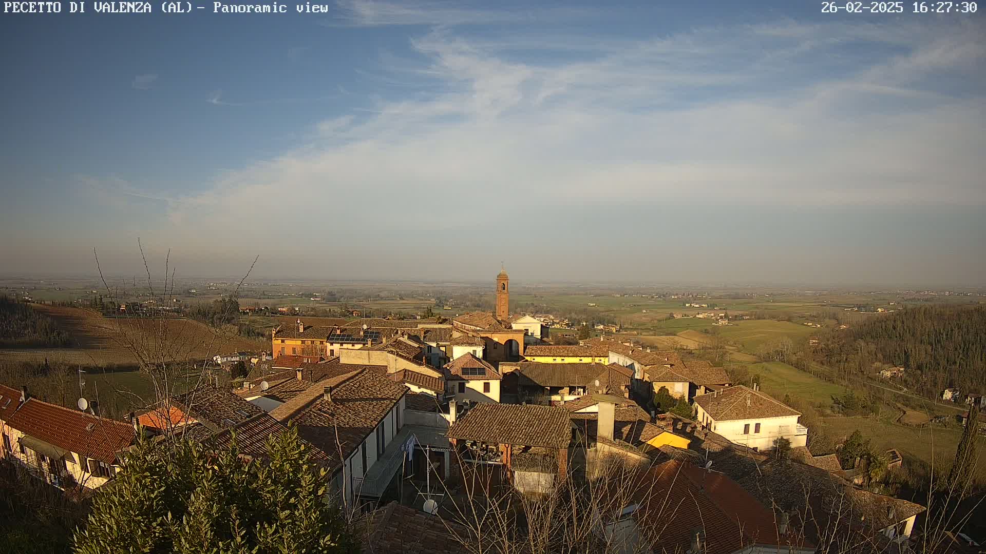 A panoramic view of a small town with terracotta roofs nestled in a valley under a mostly clear sky with a few wispy clouds.