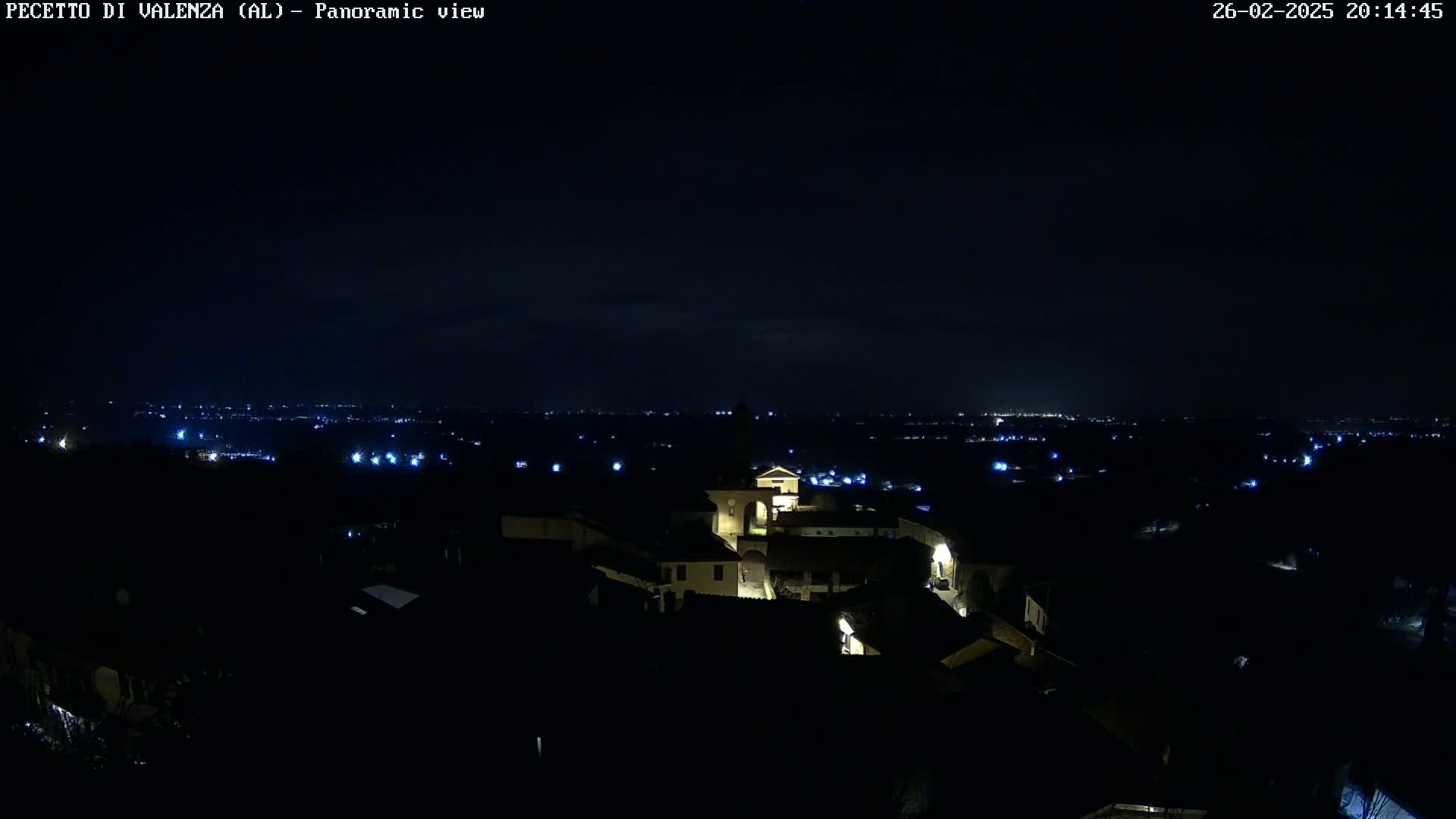 A nighttime panoramic view of a small town illuminated by scattered lights, with a darker, distant cityscape visible under a clear, dark sky.