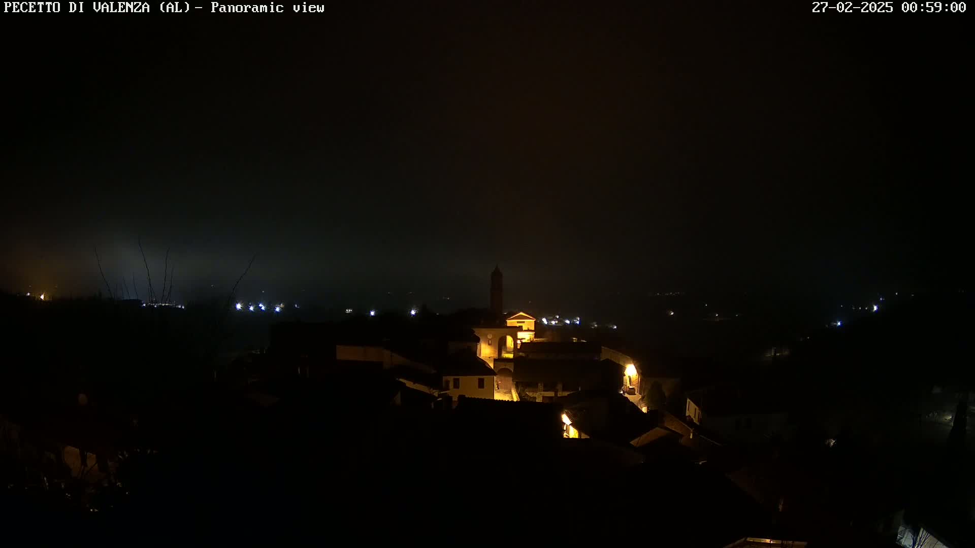 A nighttime panoramic view of a small, dimly lit town nestled in a valley under a hazy, dark sky.