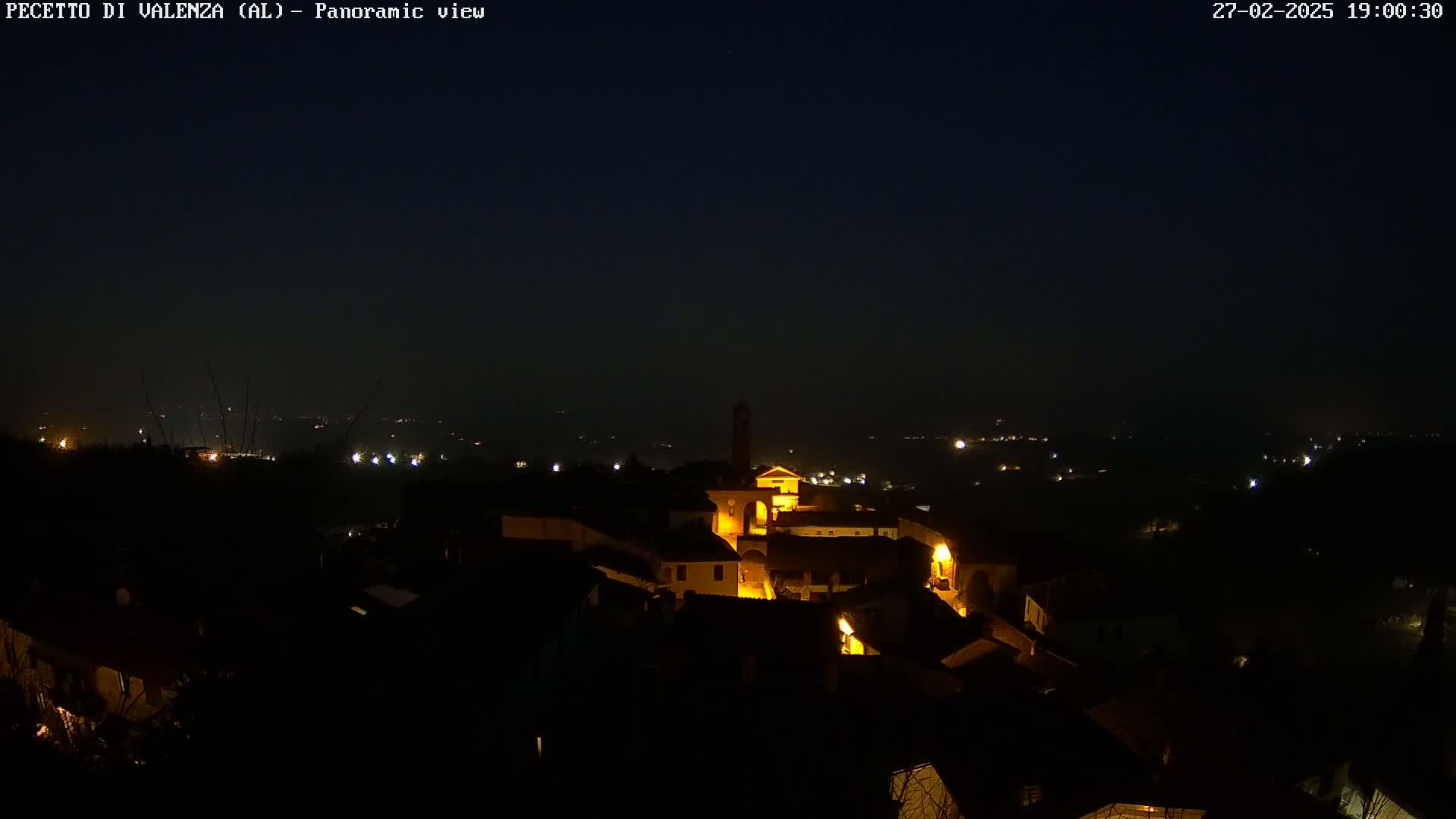 A nighttime, panoramic view of a small, illuminated town nestled in a dark, hilly landscape under a clear, dark sky.