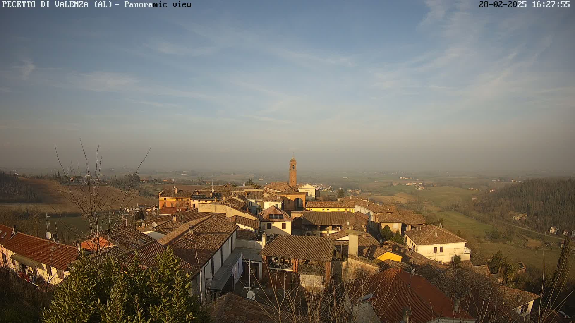 A hazy, sunny day overlooks a village of terracotta-roofed buildings nestled in rolling hills.