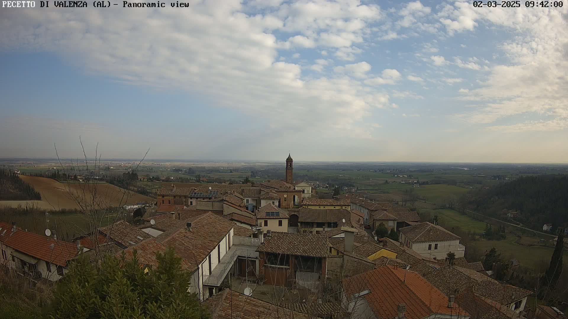 A panoramic view of a small town with red-tiled roofs, a bell tower, and a partly cloudy sky stretches over a gently rolling landscape of farmland.