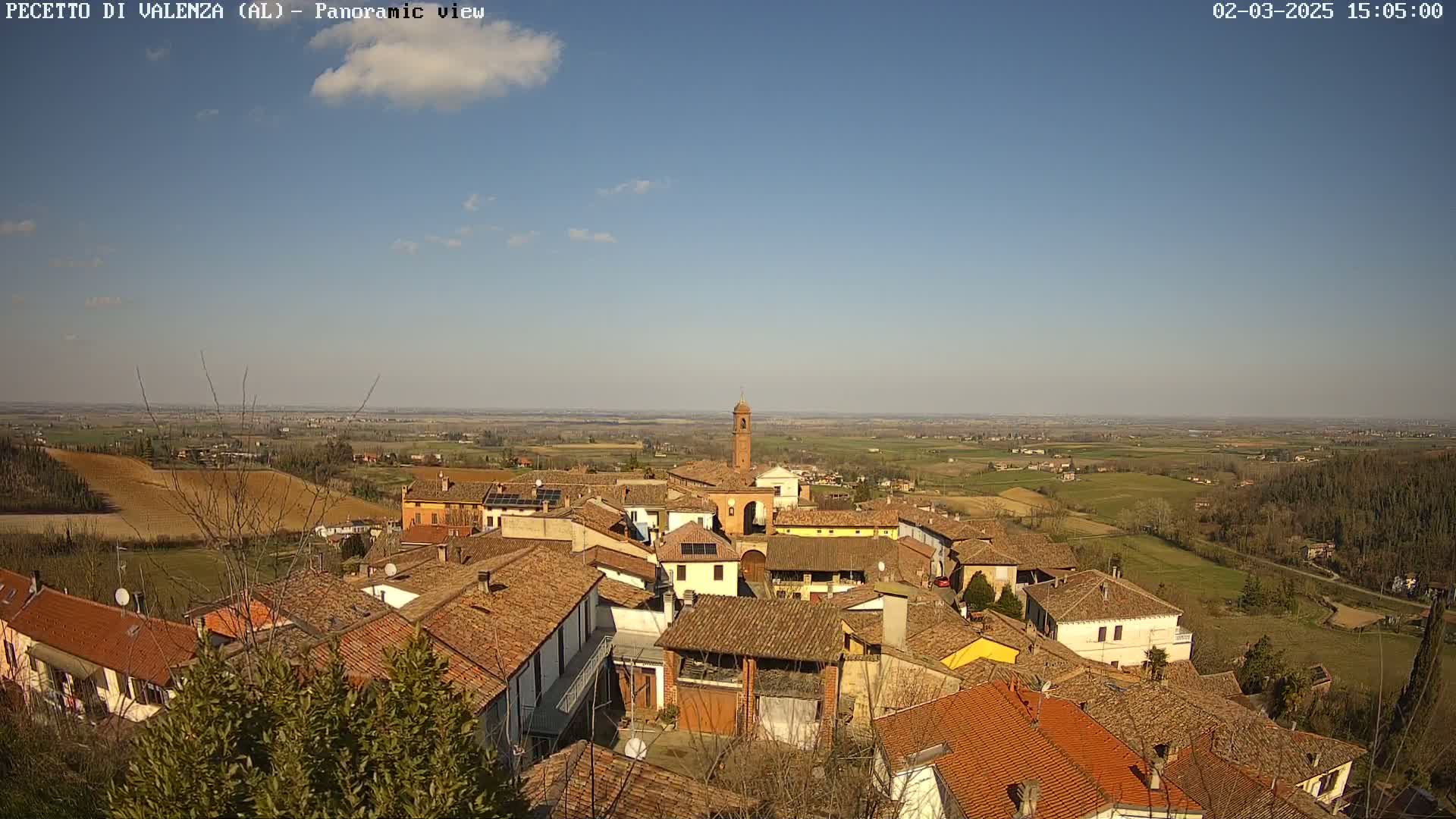 A small town with red-tiled roofs nestles on a hillside under a mostly clear blue sky, overlooking a vast, flat expanse of farmland.