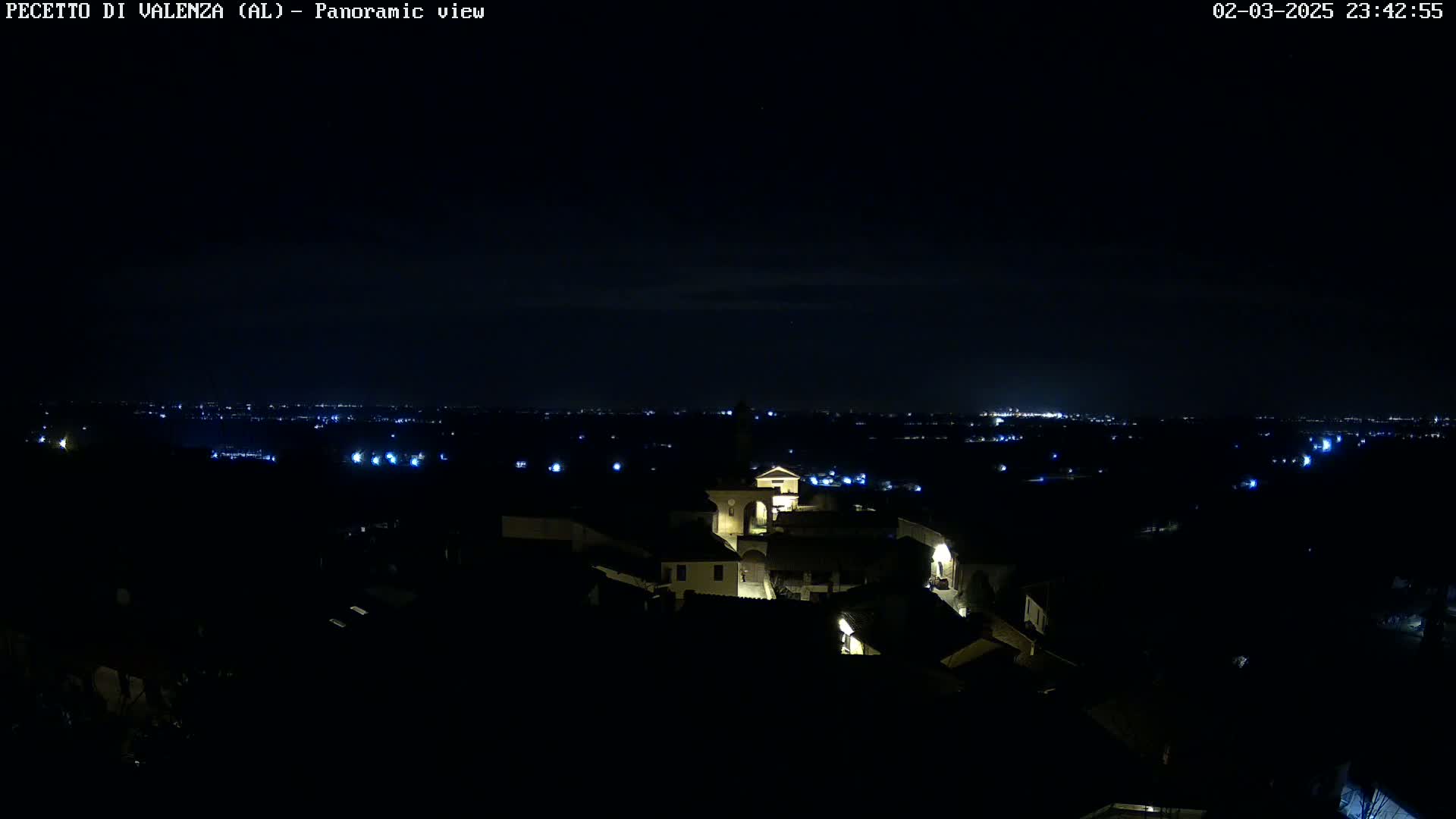A nighttime panoramic view of a small town with dimly lit buildings, surrounded by a dark landscape with sparsely lit areas in the distance under a clear, dark sky.
