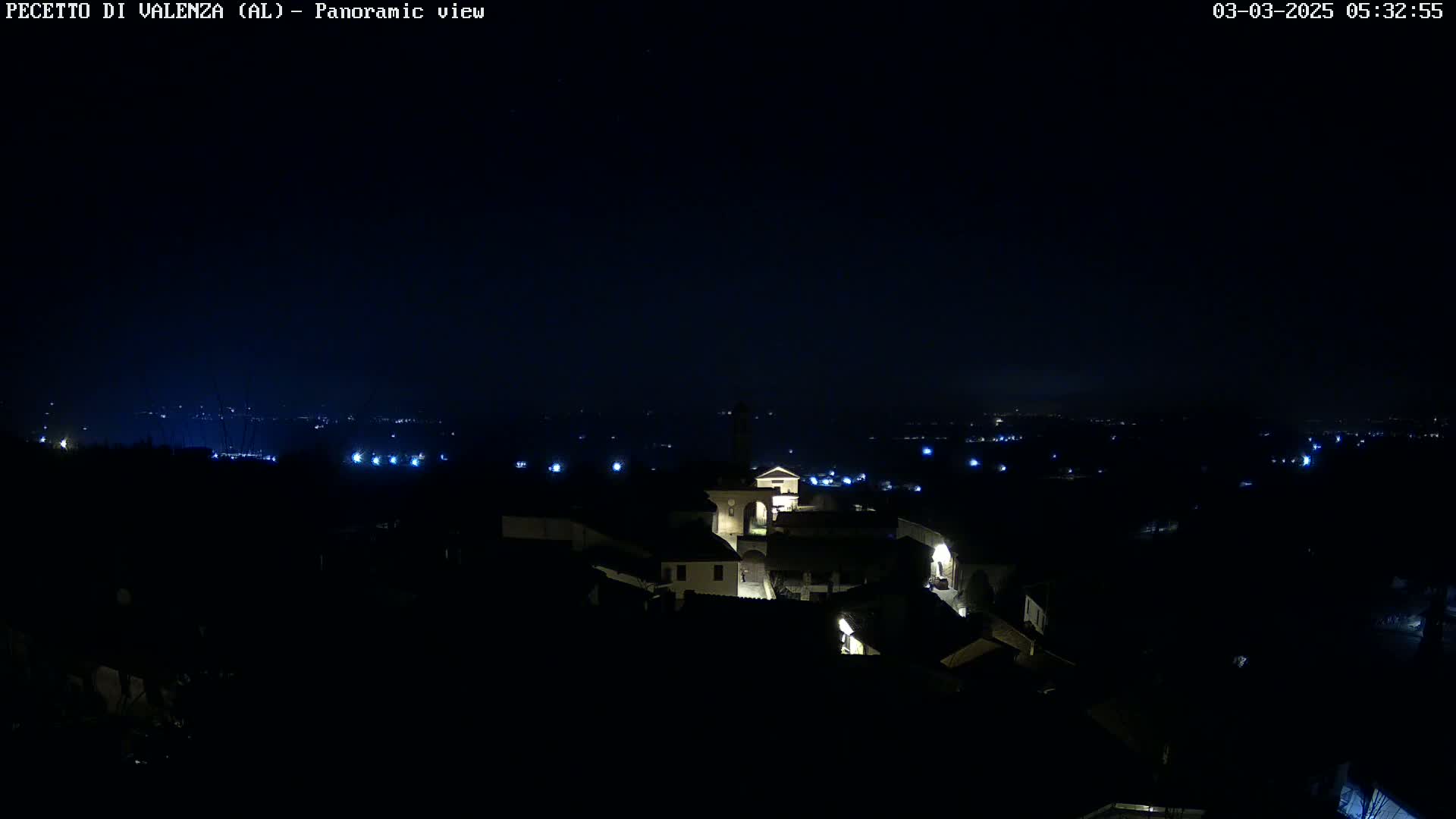 A nighttime panoramic view of a small, dimly lit town nestled in a dark landscape under a clear, starless sky.