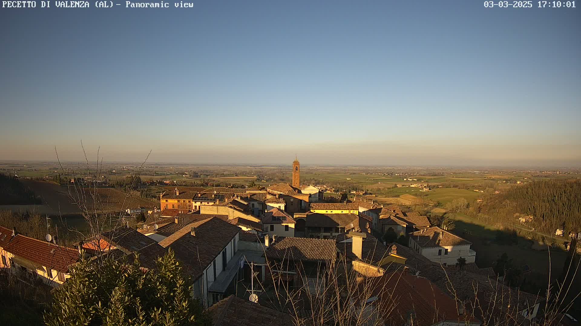 A panoramic view of a small town with red-tiled roofs under a clear, late afternoon sky, showing surrounding farmland extending to the horizon.