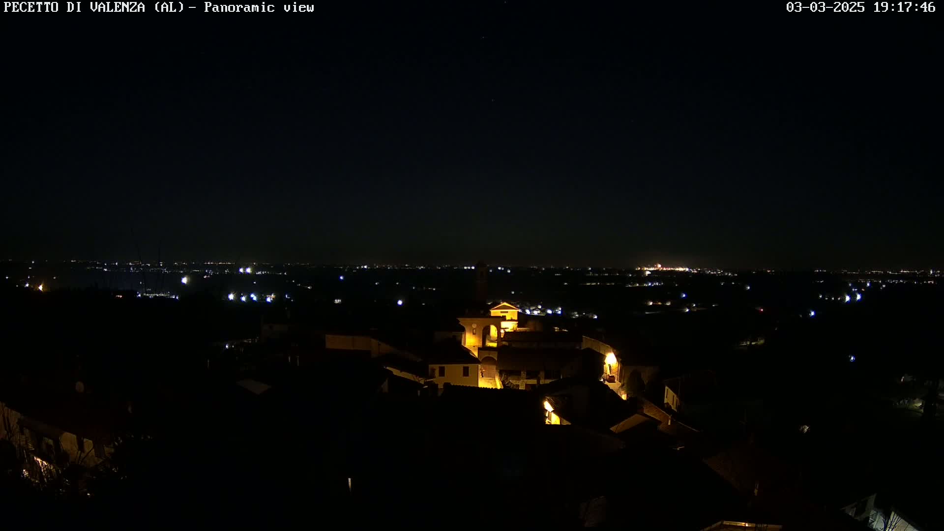 A nighttime panoramic view shows a small, illuminated village nestled in a dark landscape, with a larger city's lights visible in the distance under a clear, dark sky.