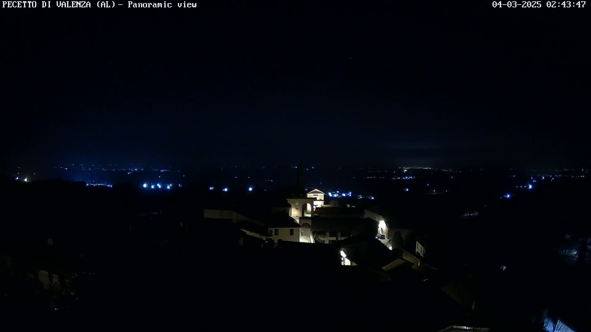 A nighttime panoramic view shows a small, dimly lit town nestled in a dark landscape under a clear, starless sky.