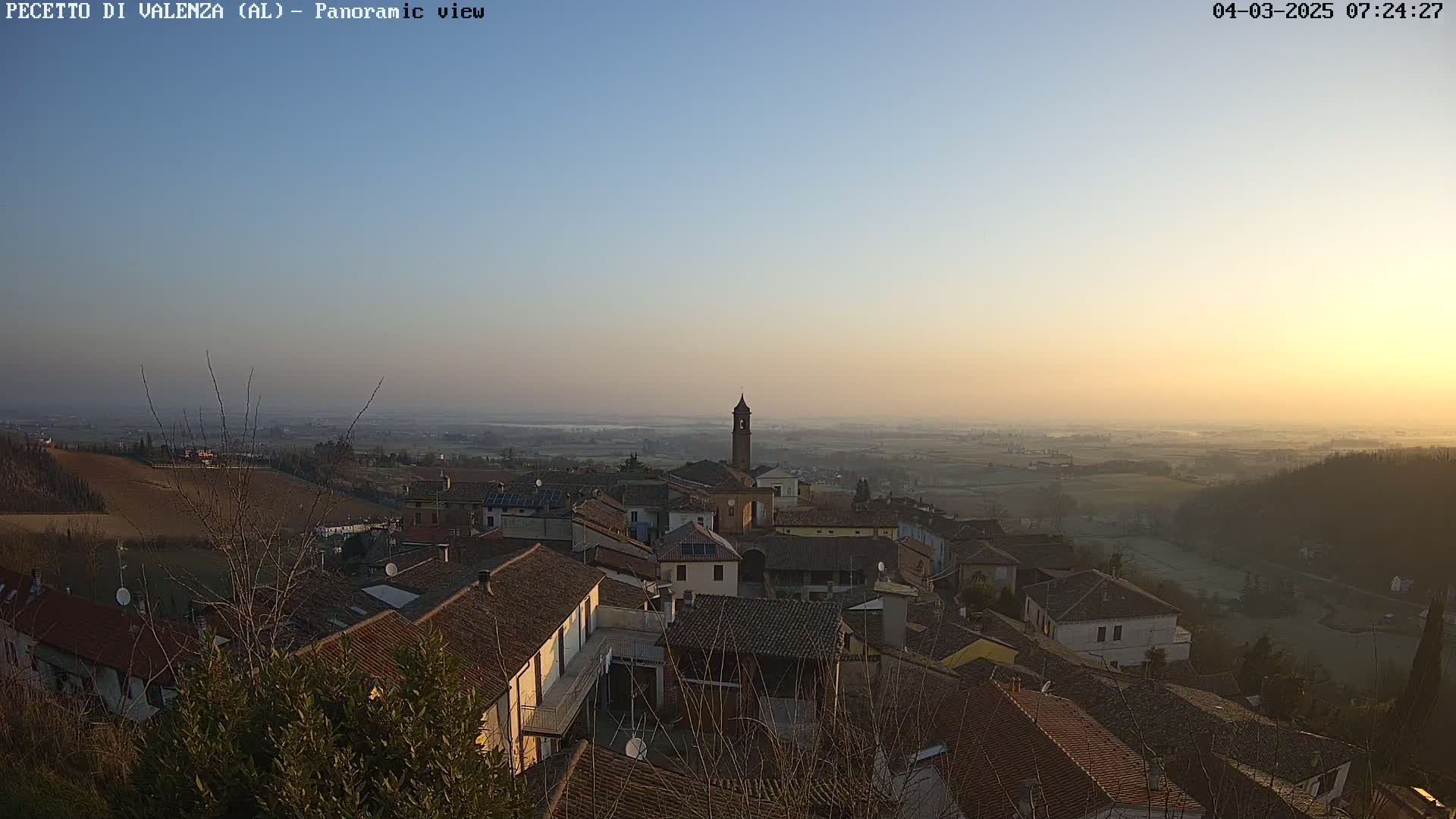 A hazy sunrise illuminates a small town nestled in a valley, with rolling hills and farmland stretching into the distance.