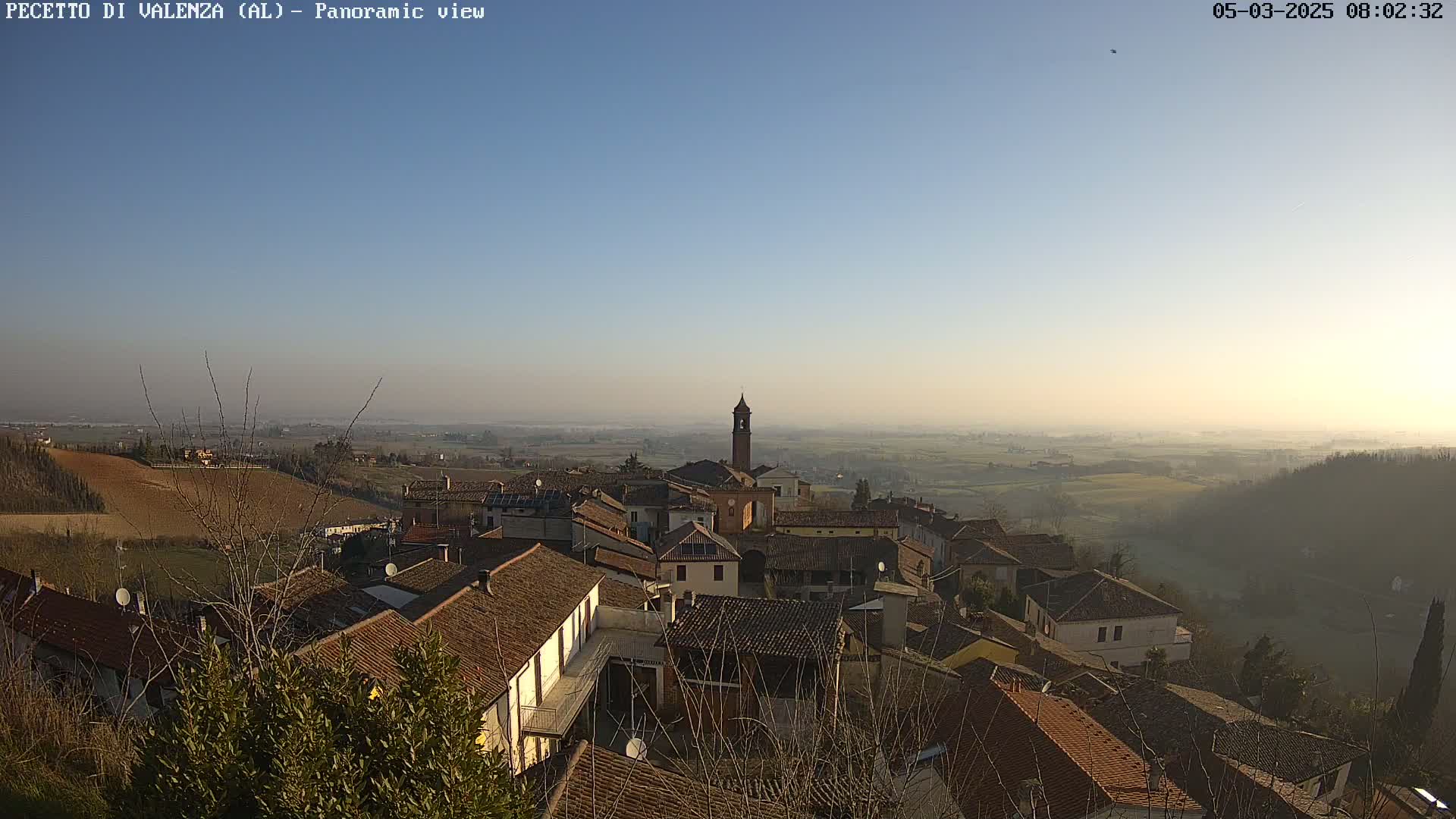 A hazy, sunny day reveals a panoramic view of a small village nestled in a hilly landscape, with a tall church tower as a prominent feature.