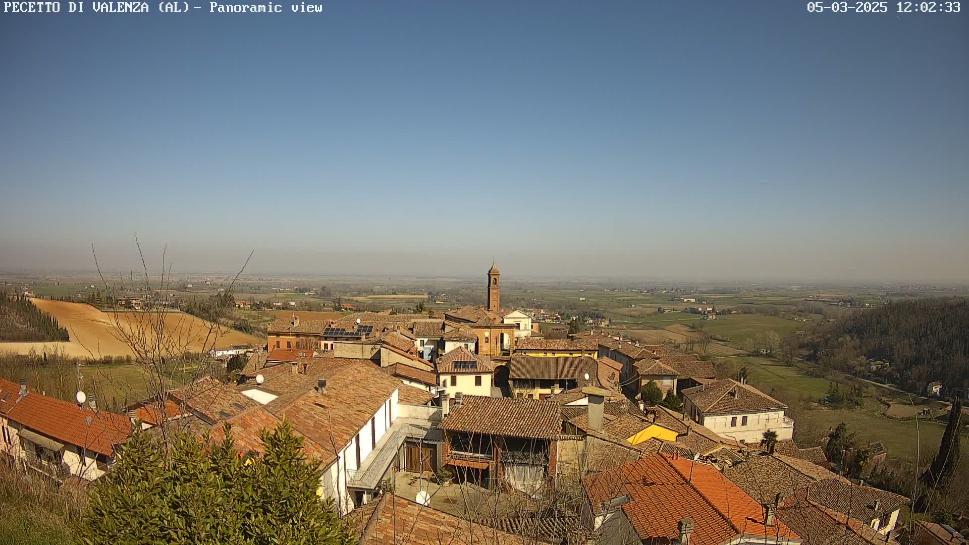A panoramic view of a small town nestled in a hilly landscape under a clear blue sky shows terracotta rooftops and a bell tower.