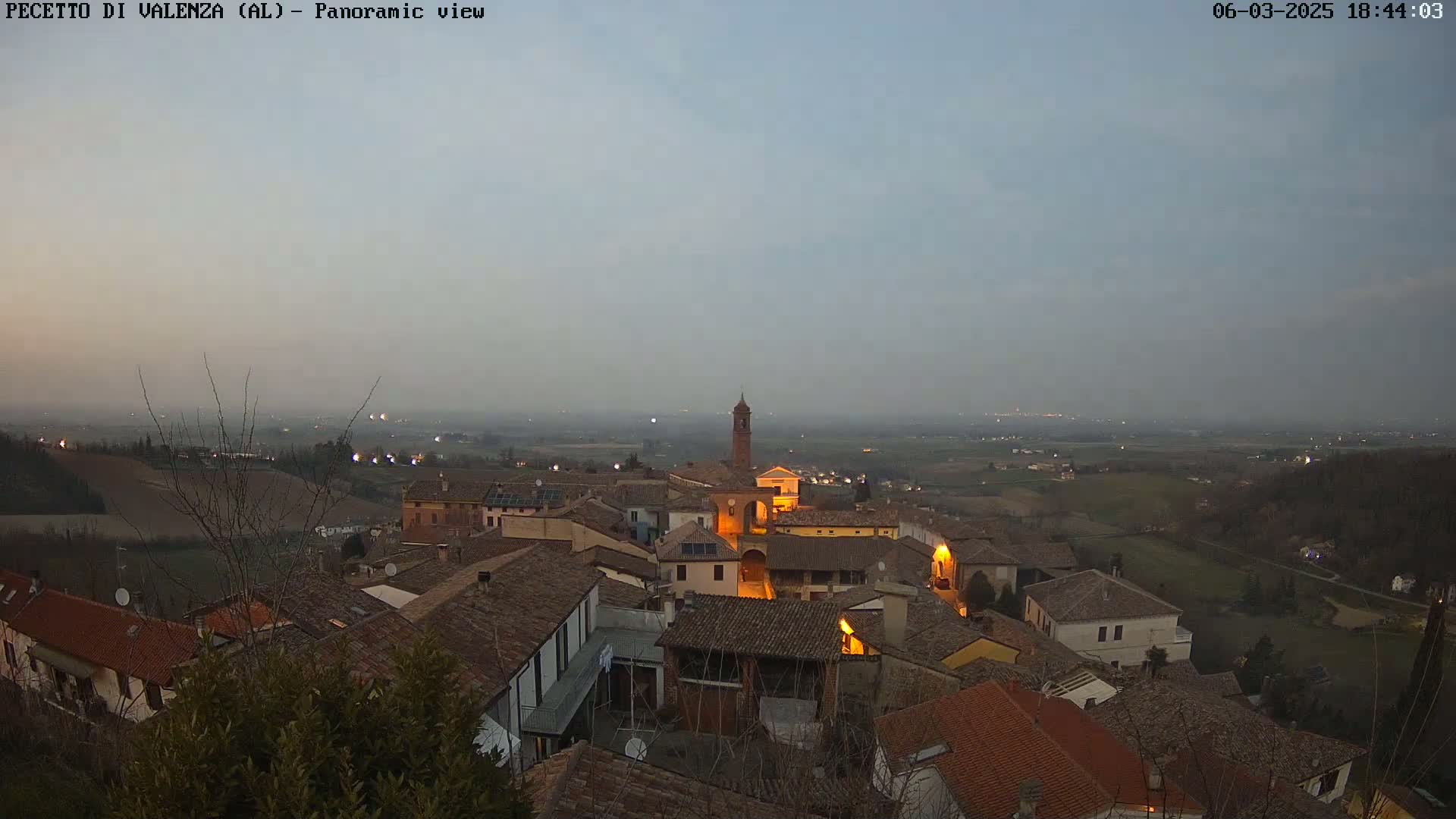 A hazy twilight view overlooks a small village nestled in a hilly landscape, with a few lights illuminating the buildings.