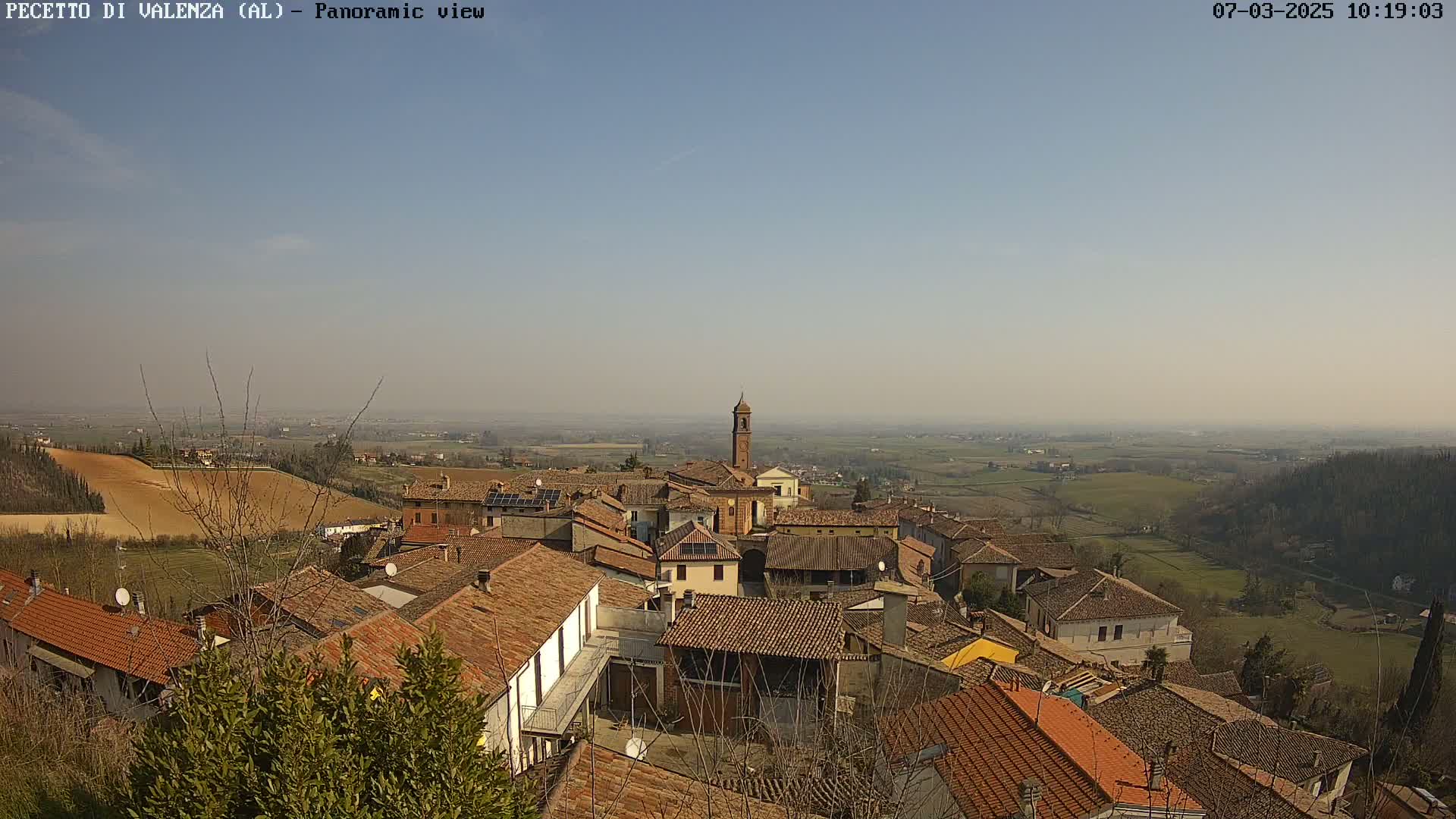 A hazy, sunny day overlooks a village of red-tiled roofs and a central bell tower, situated on a hillside overlooking farmland.