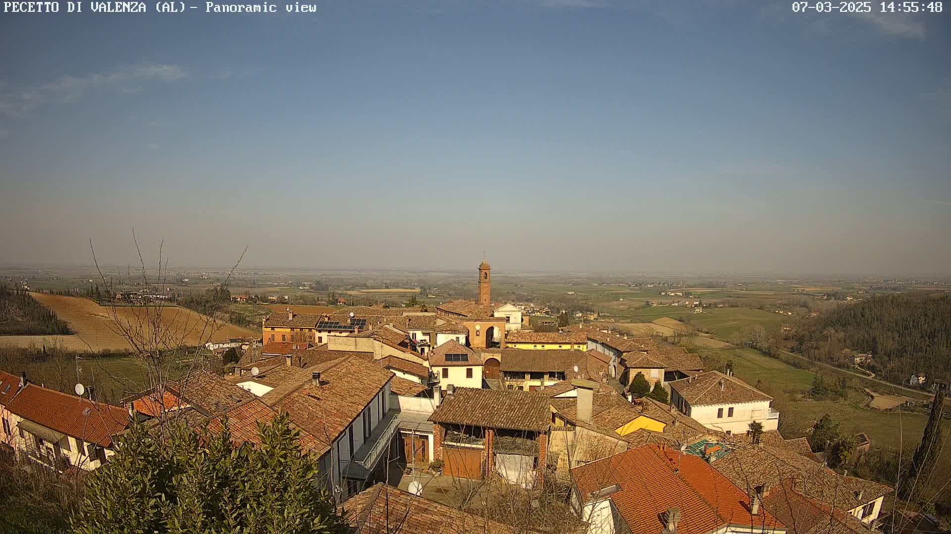 A panoramic view of a small village with red-tiled roofs, a tall bell tower, and surrounding farmland under a clear, sunny sky.