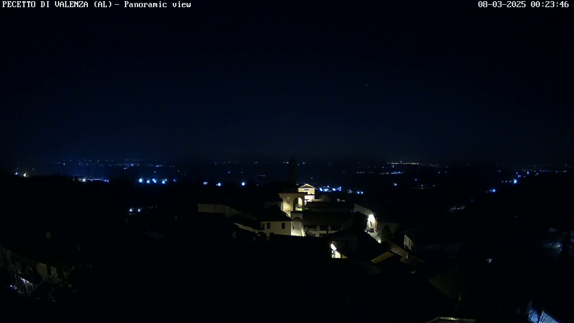 A nighttime panoramic view of a small, dimly lit village nestled on a hilltop, overlooking a distant expanse of lights under a dark, clear sky.
