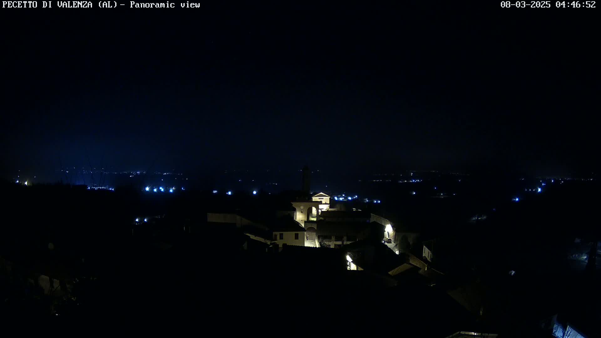 A nighttime panoramic view shows a small, dimly lit village nestled in a dark landscape under a clear, starless sky.