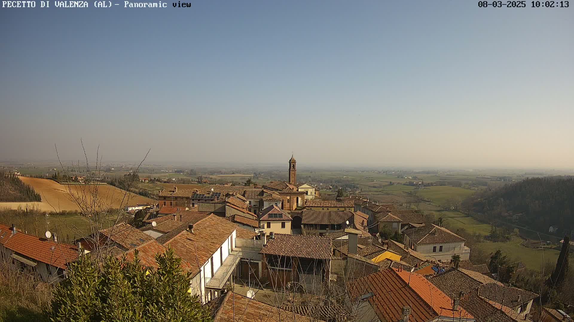 A panoramic view of a small town with terracotta rooftops, set against a hazy, sunny day and a flat, agricultural landscape.