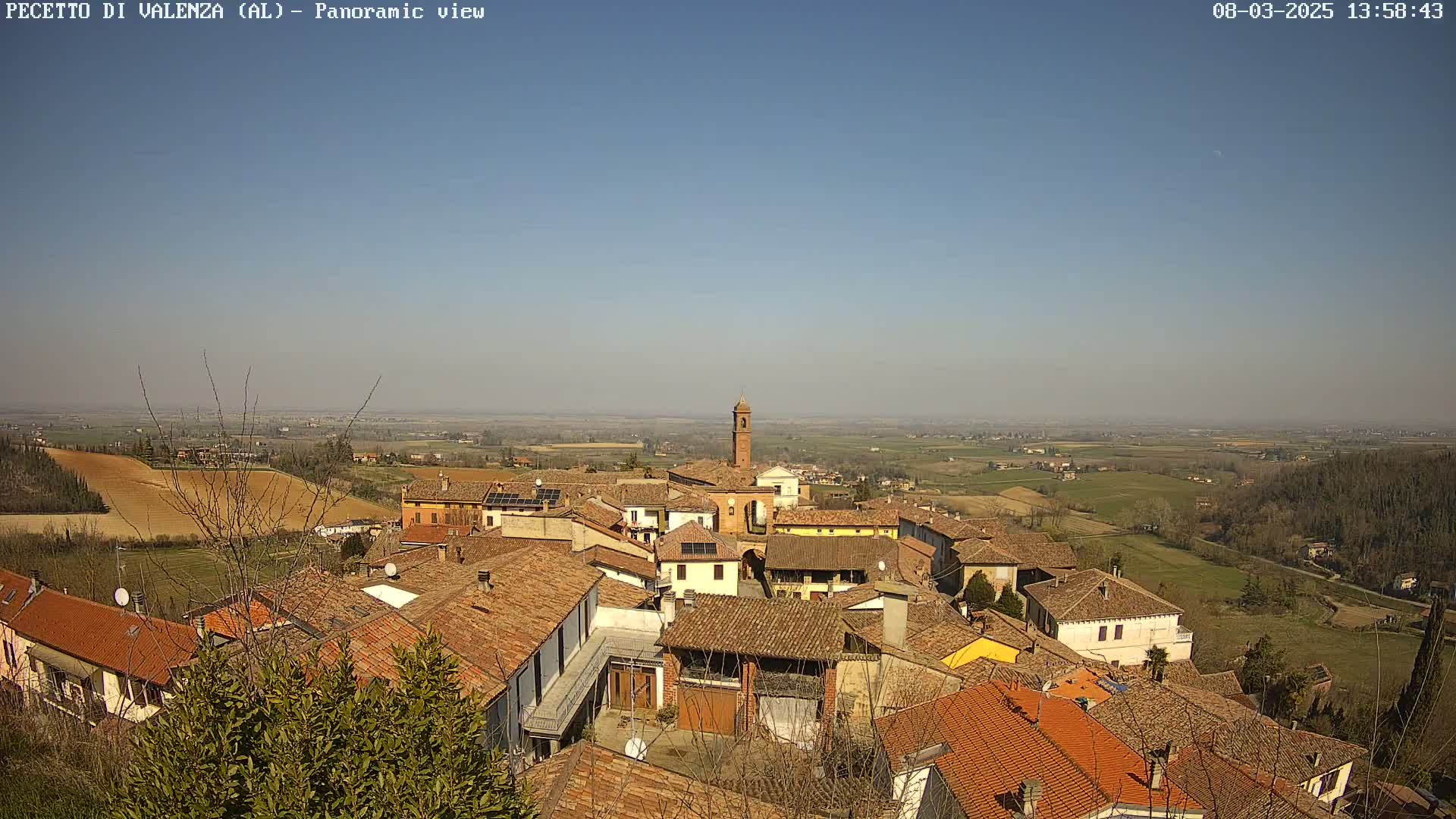 A panoramic view of a small town nestled in a valley on a clear, sunny day, with terracotta-roofed buildings and a distant expanse of farmland.