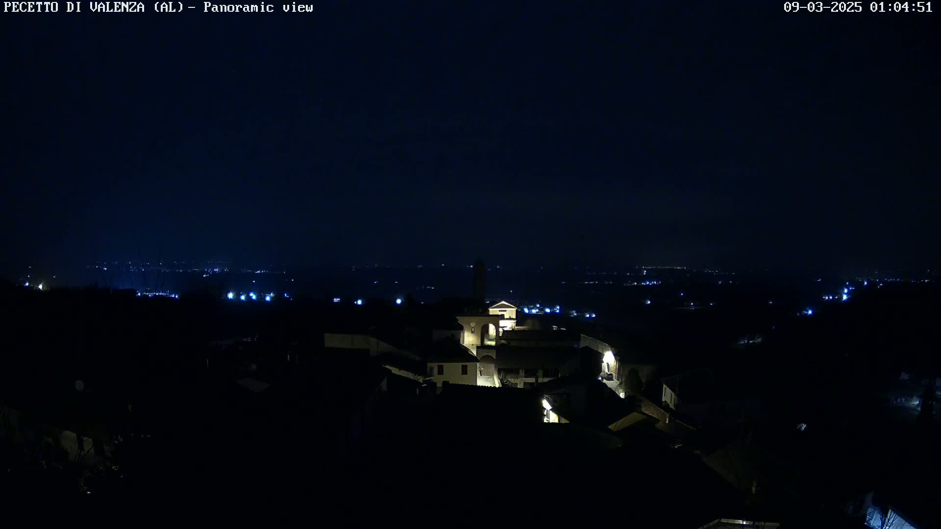 A small, dark village is illuminated by scattered interior lights at night, overlooking a distant, dimly lit landscape under a clear, dark sky.