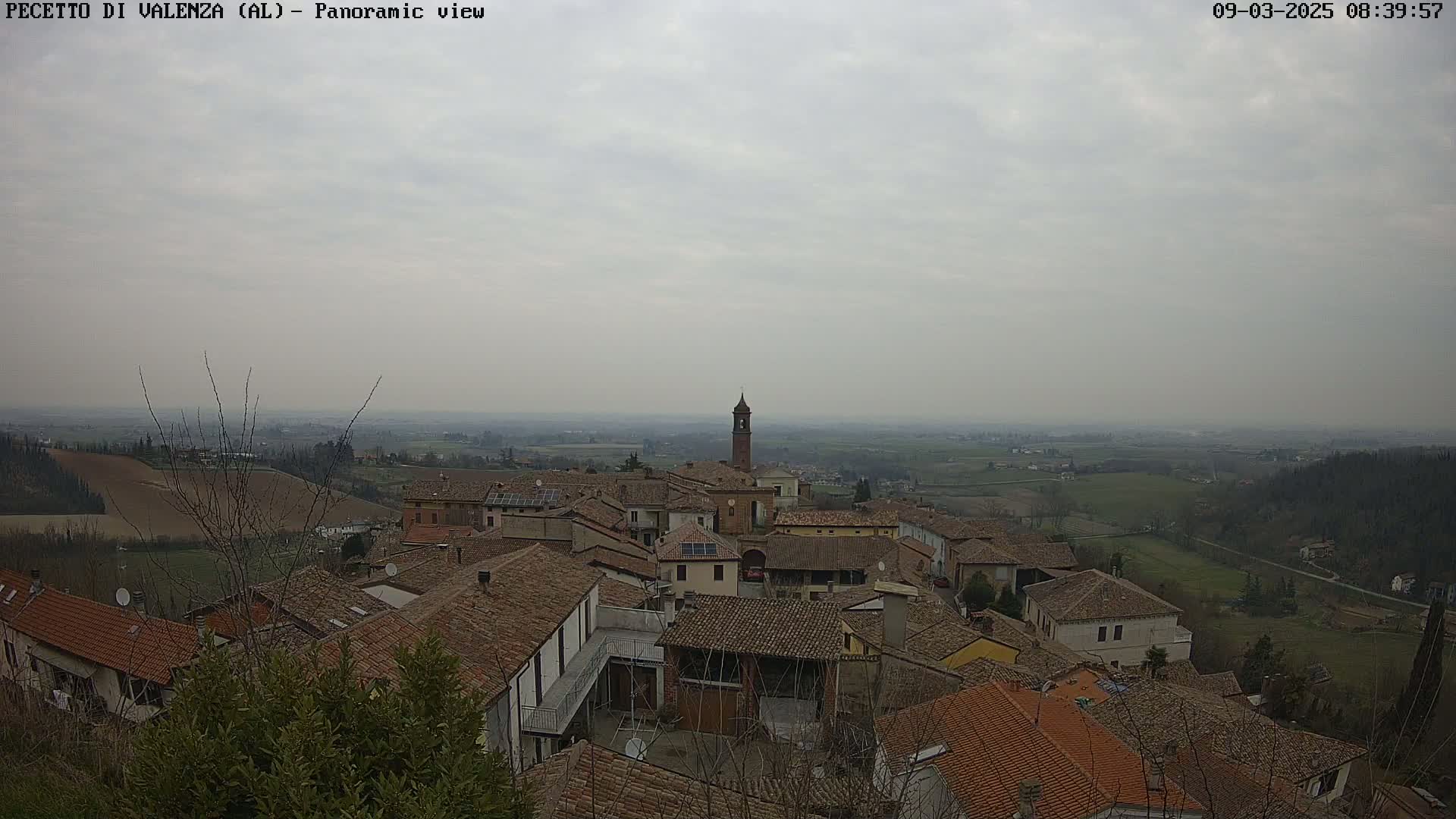 A panoramic view of a small town with red-tiled roofs under an overcast sky, set against a hazy landscape of rolling hills and farmland.