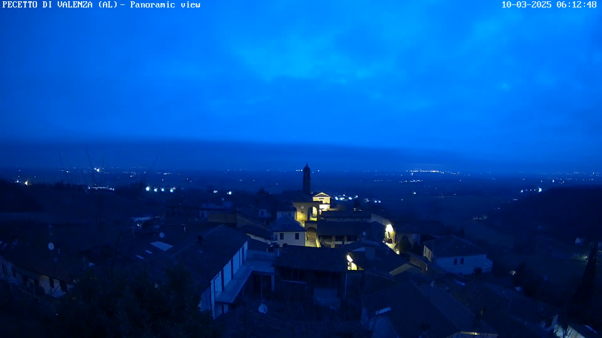 A nighttime panoramic view of a small town illuminated by a few lights, under a dark blue, mostly clear sky.