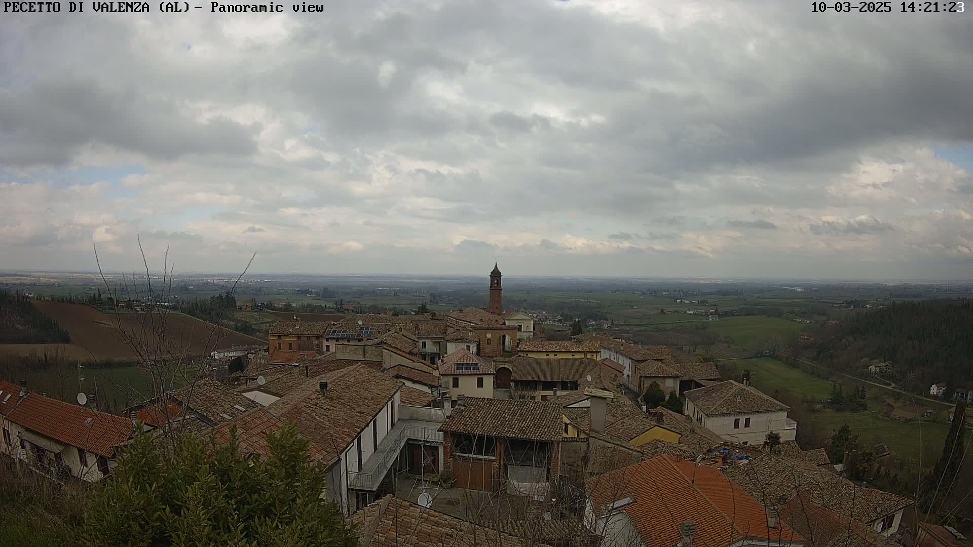 A panoramic view of a town with red-tiled roofs under a cloudy sky, overlooking a flat, green landscape.