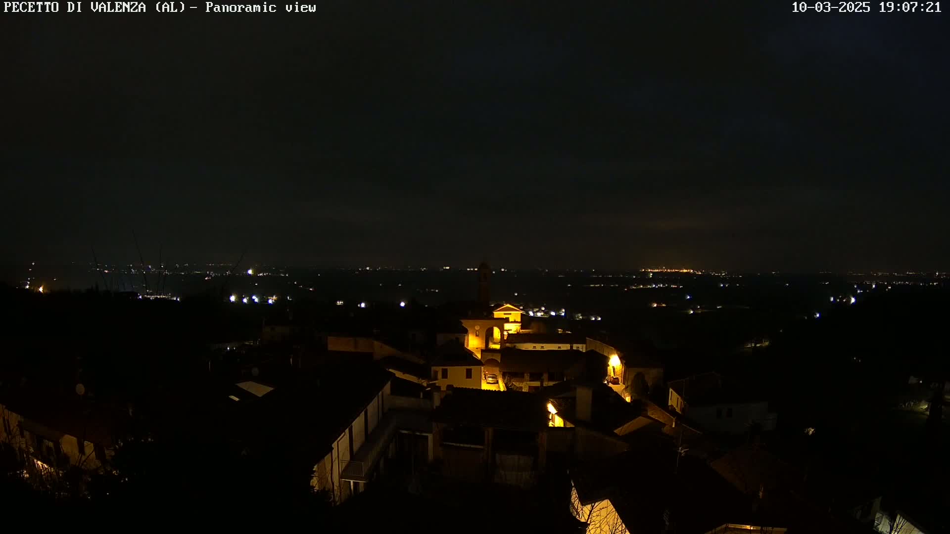 A nighttime panoramic view of a small, illuminated town nestled in a dark, hilly landscape under a mostly clear sky.