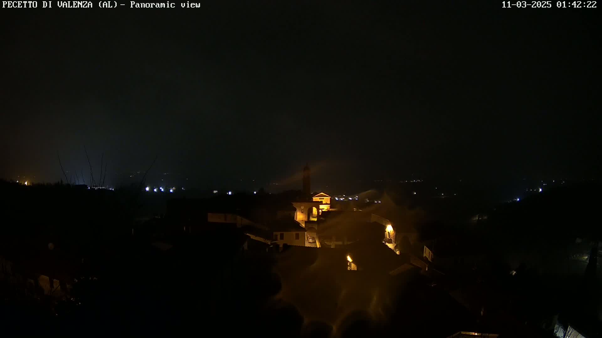 A nighttime panoramic view of a small village with dimly lit buildings, under a dark, possibly foggy or hazy sky.