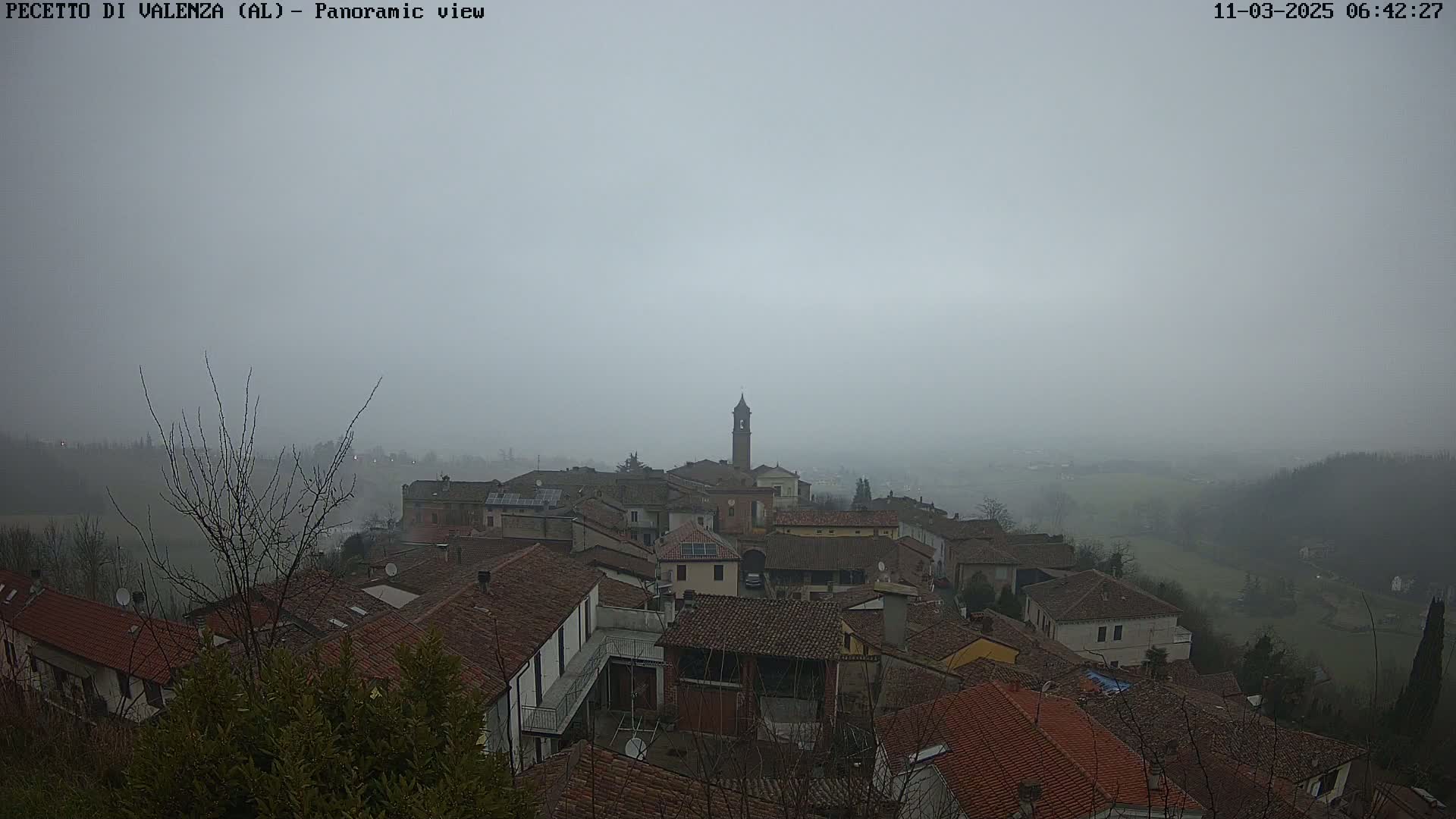 A foggy, overcast day reveals a panoramic view of a village with red-tiled roofs and a tall bell tower, nestled in a valley surrounded by hills.