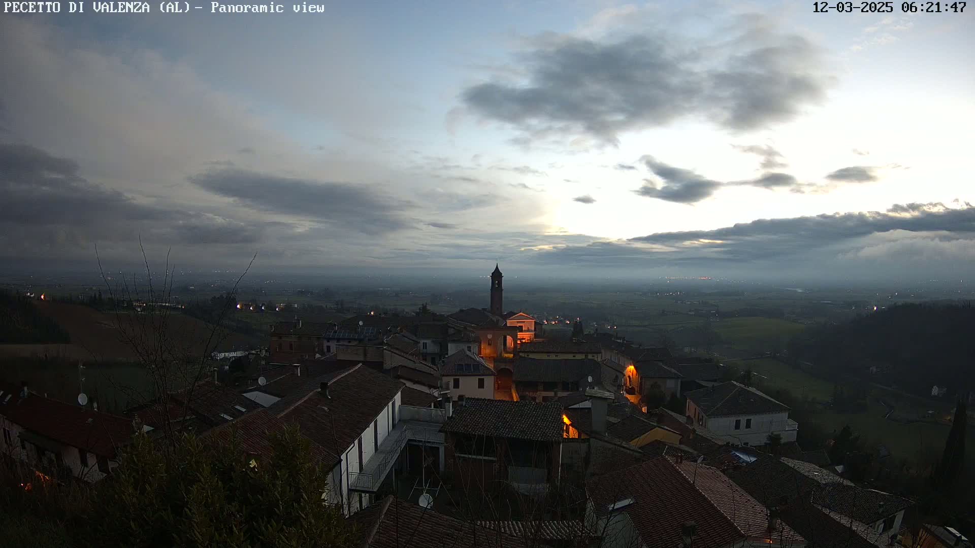 A small town with dimly lit buildings sits on a hilltop under a cloudy, pre-dawn sky.