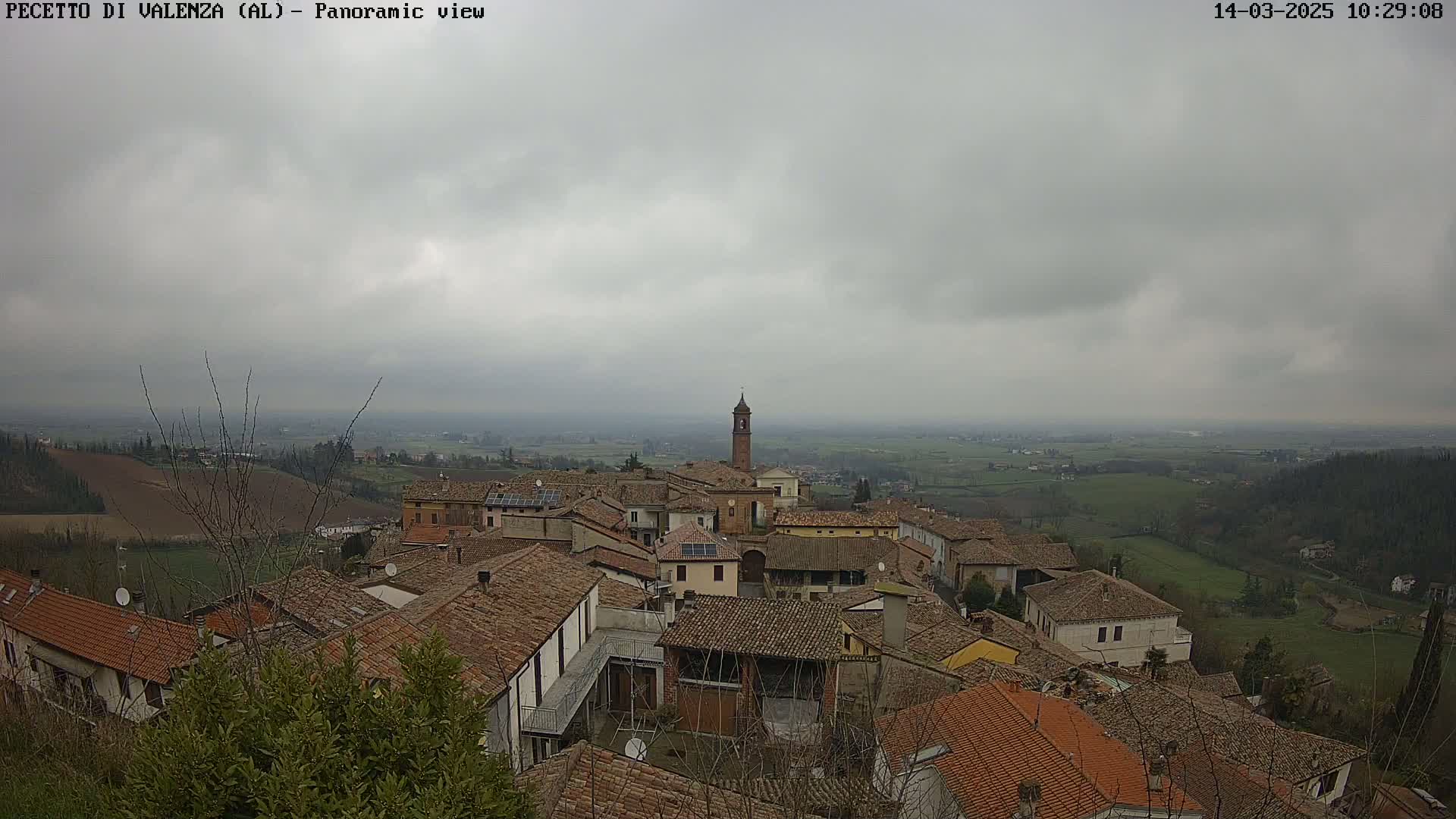 A panoramic view of a town with terracotta roofs under an overcast sky shows a valley stretching into the distance.